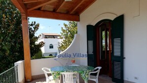Un'area esterna coperta con un tavolo e sedie, un muro bianco e una porta con persiane verdi.