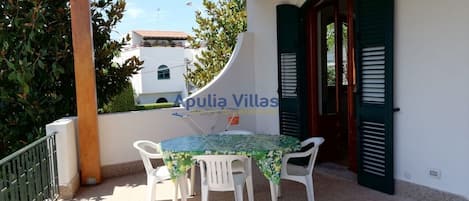 A covered outdoor area with a table and chairs, a white wall, and a door with green shutters.