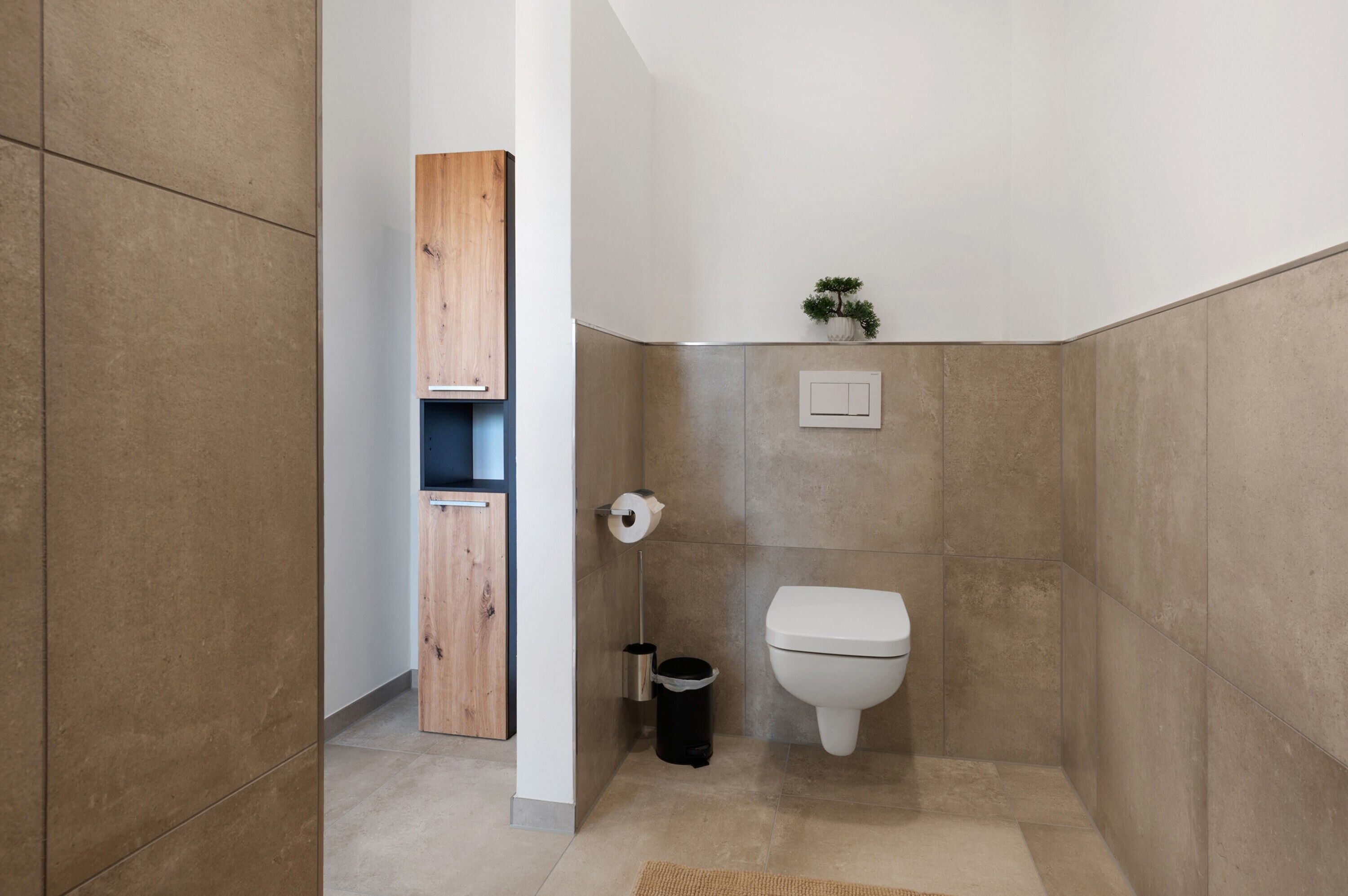 A bathroom with a toilet, a trash can, a wooden cabinet, and a small plant on the wall.