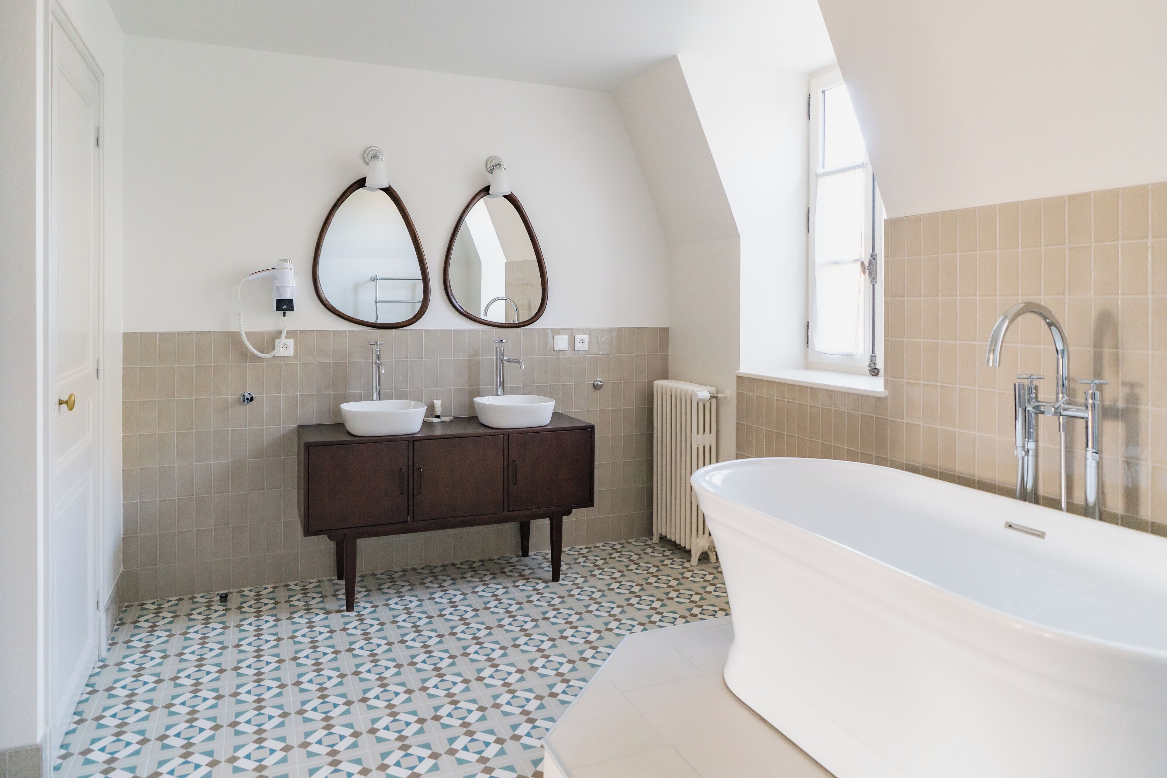 Une salle de bain moderne avec une baignoire sur pieds, deux vasques avec des miroirs ronds et un carrelage au motif décoratif.