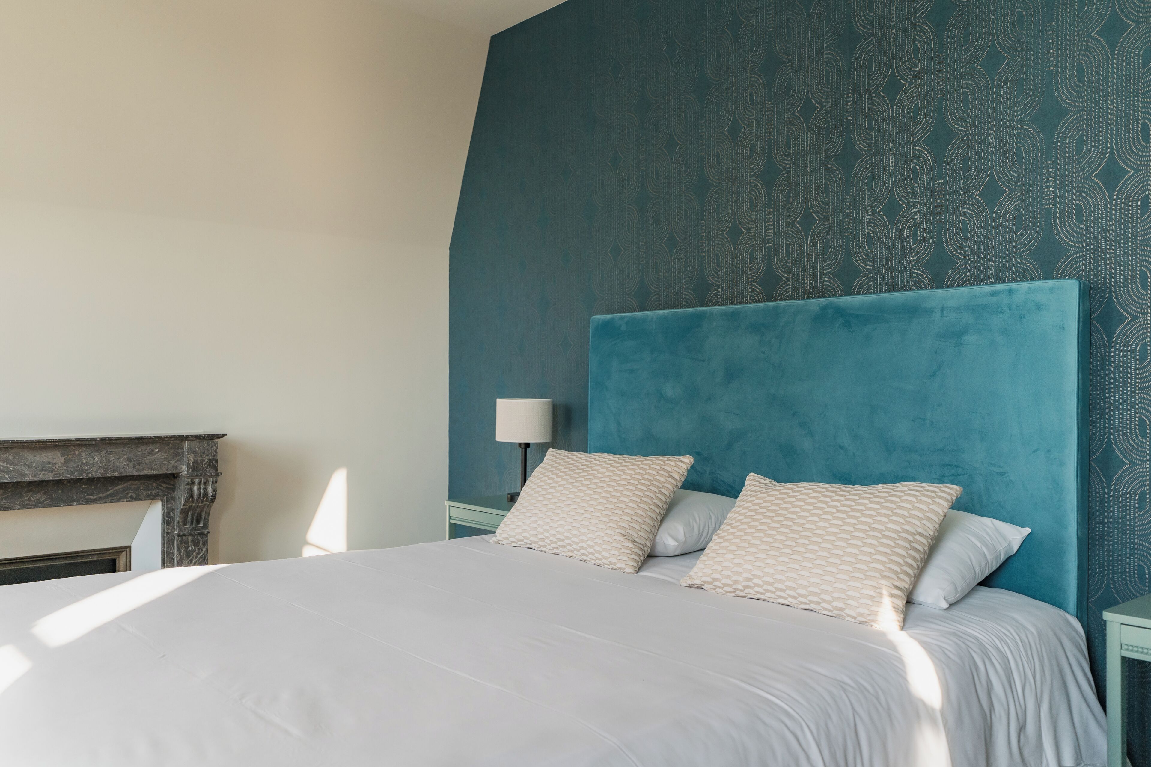 A neatly made bed with a white duvet and patterned pillows, a blue headboard, and a patterned wall.