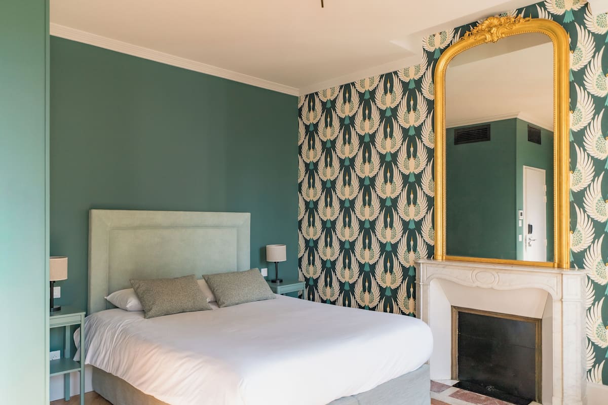 A bedroom with a large bed, a nightstand, a lamp, a fireplace, and a mirror with a gold frame.