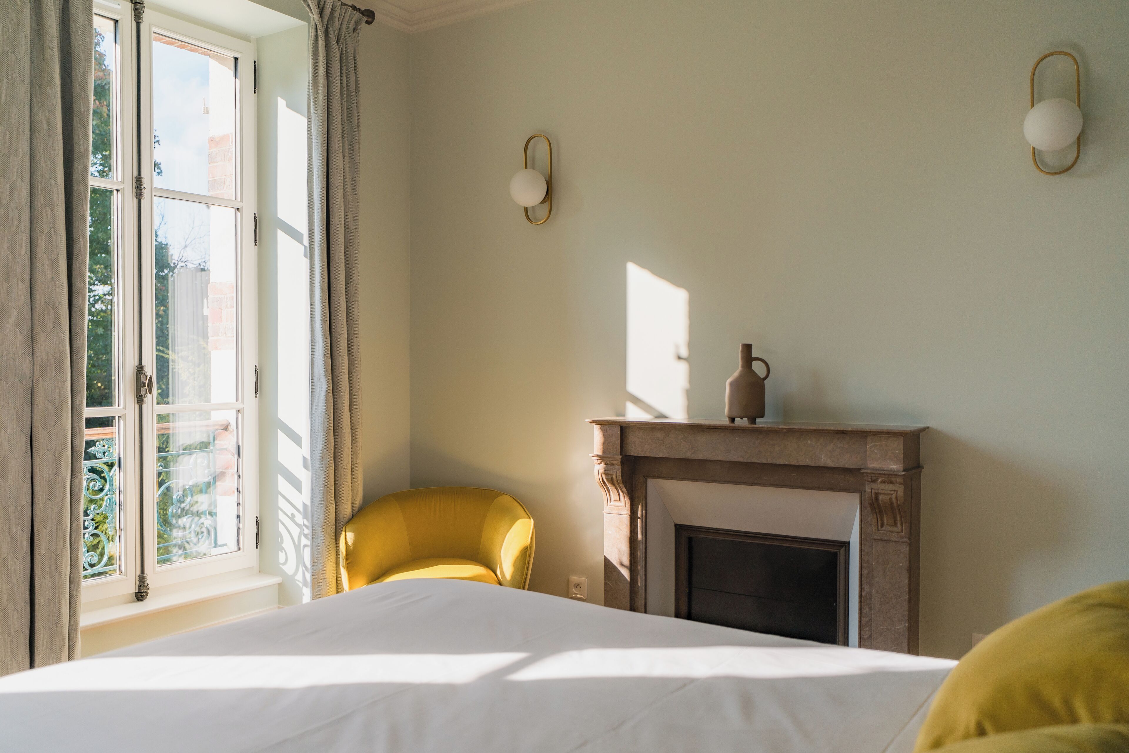 A bedroom with a bed, a yellow armchair, a fireplace, and a window with curtains.