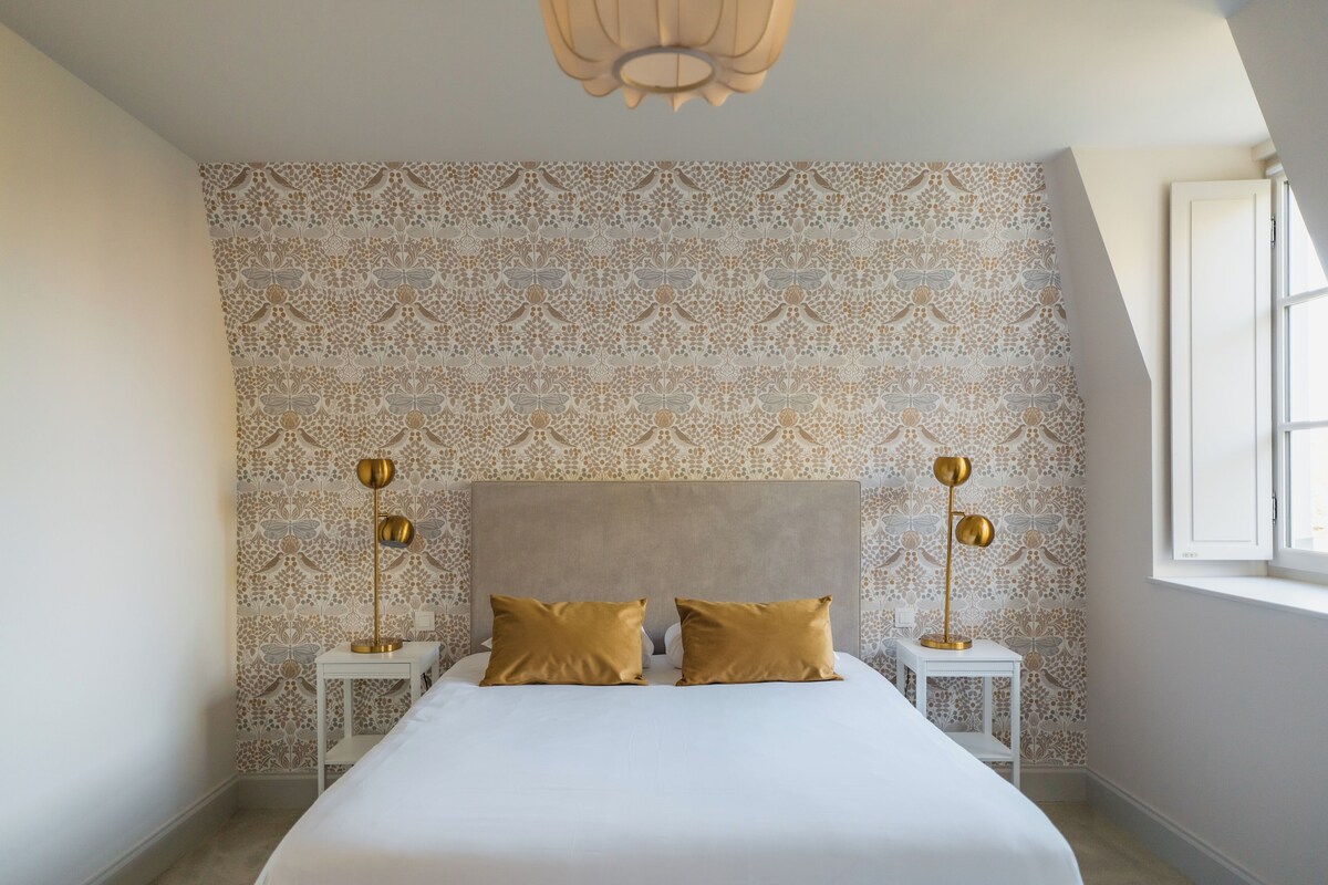 A bedroom with a patterned wallpaper, a large bed with two gold pillows, and two bedside lamps.