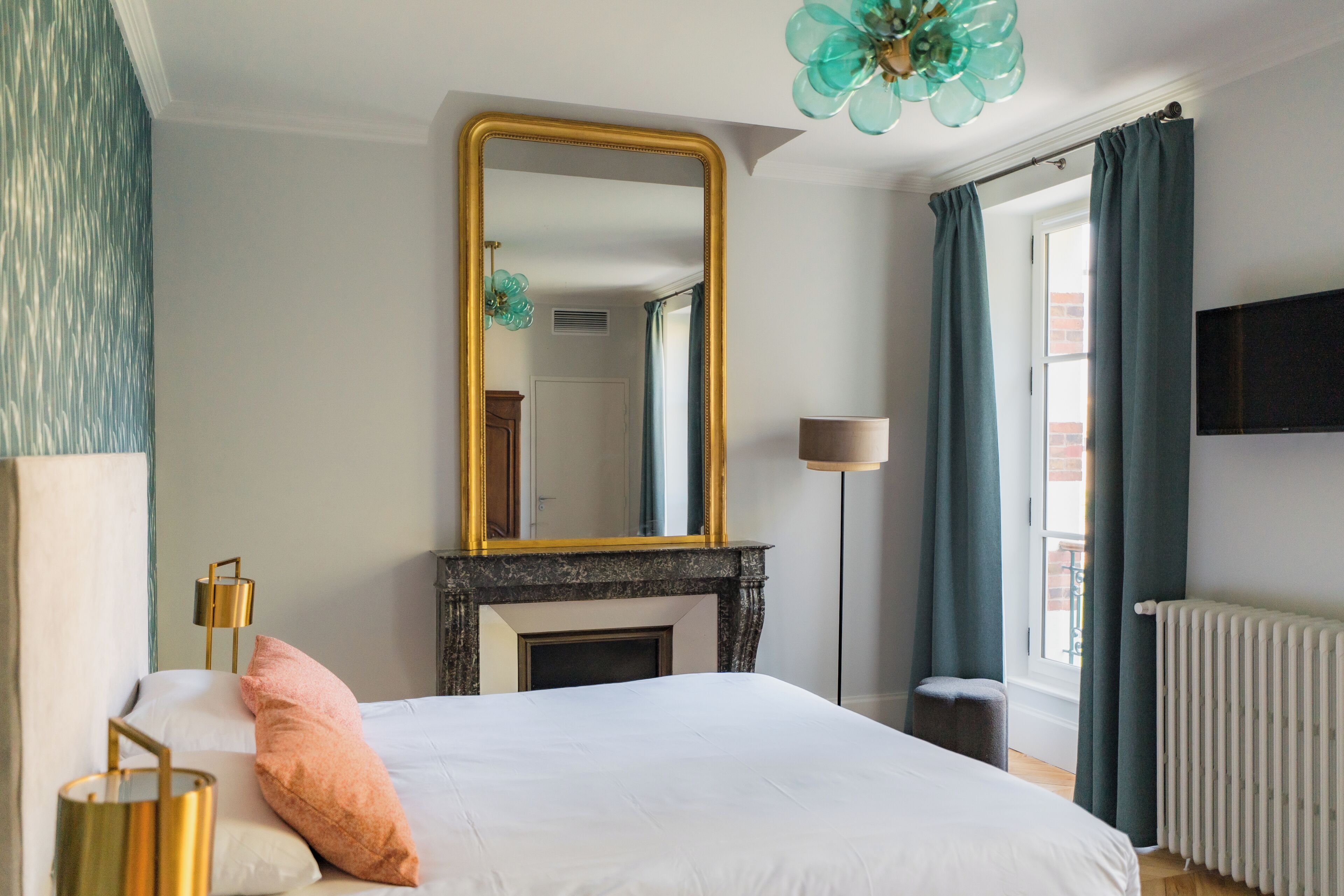 A bedroom with a bed, a large mirror, a fireplace, a window with curtains, and a radiator.