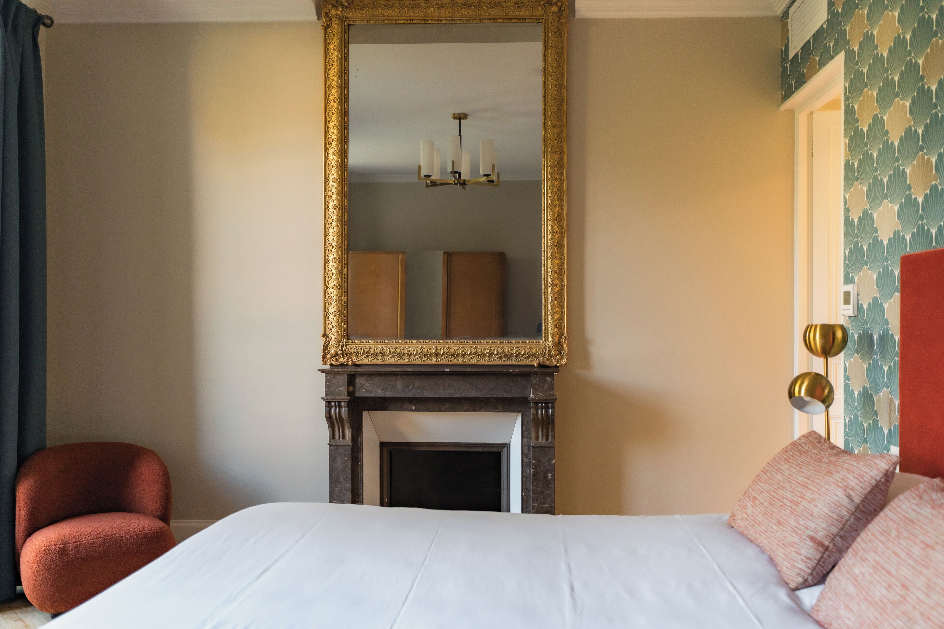 A bedroom with a large bed, a fireplace, a mirror, and a chair.