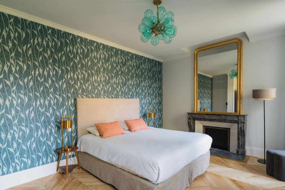 A bedroom with a large bed, a fireplace, a mirror, and patterned wallpaper.