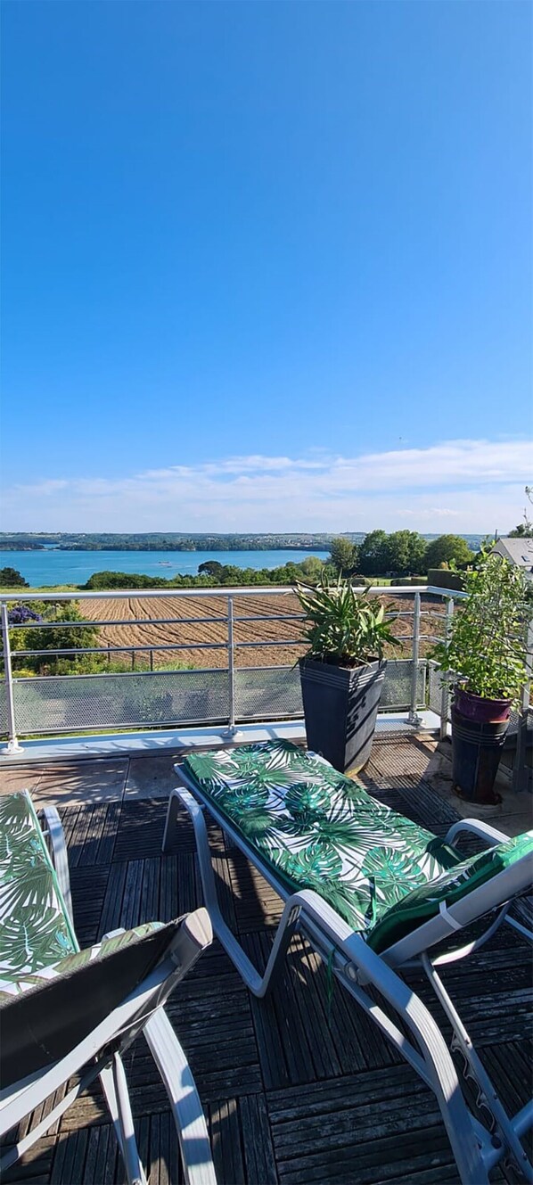 A deck with outdoor furniture and potted plants, overlooking a body of water and fields.