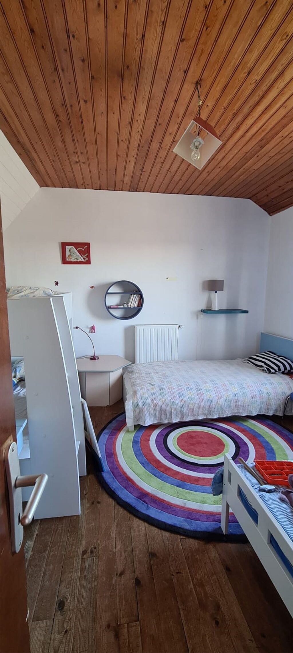 A small, cozy room with wooden ceiling beams, a bed with a rainbow-patterned rug, a desk with a lamp, and a radiator.