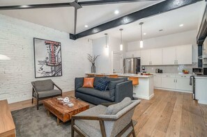 A modern living room with a sofa, armchairs, and a coffee table. A kitchen with white cabinets and stainless steel appliances is visible in the background.