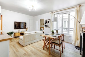 A modern living room with a sofa, a dining table with chairs, a TV mounted on the wall, and a large window with curtains.