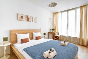 A hotel room with a large bed, a bedside table with a lamp, a small table with a plant, and a window with curtains.