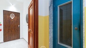Um corredor com uma porta de madeira, uma porta azul com janela e uma parede amarela.