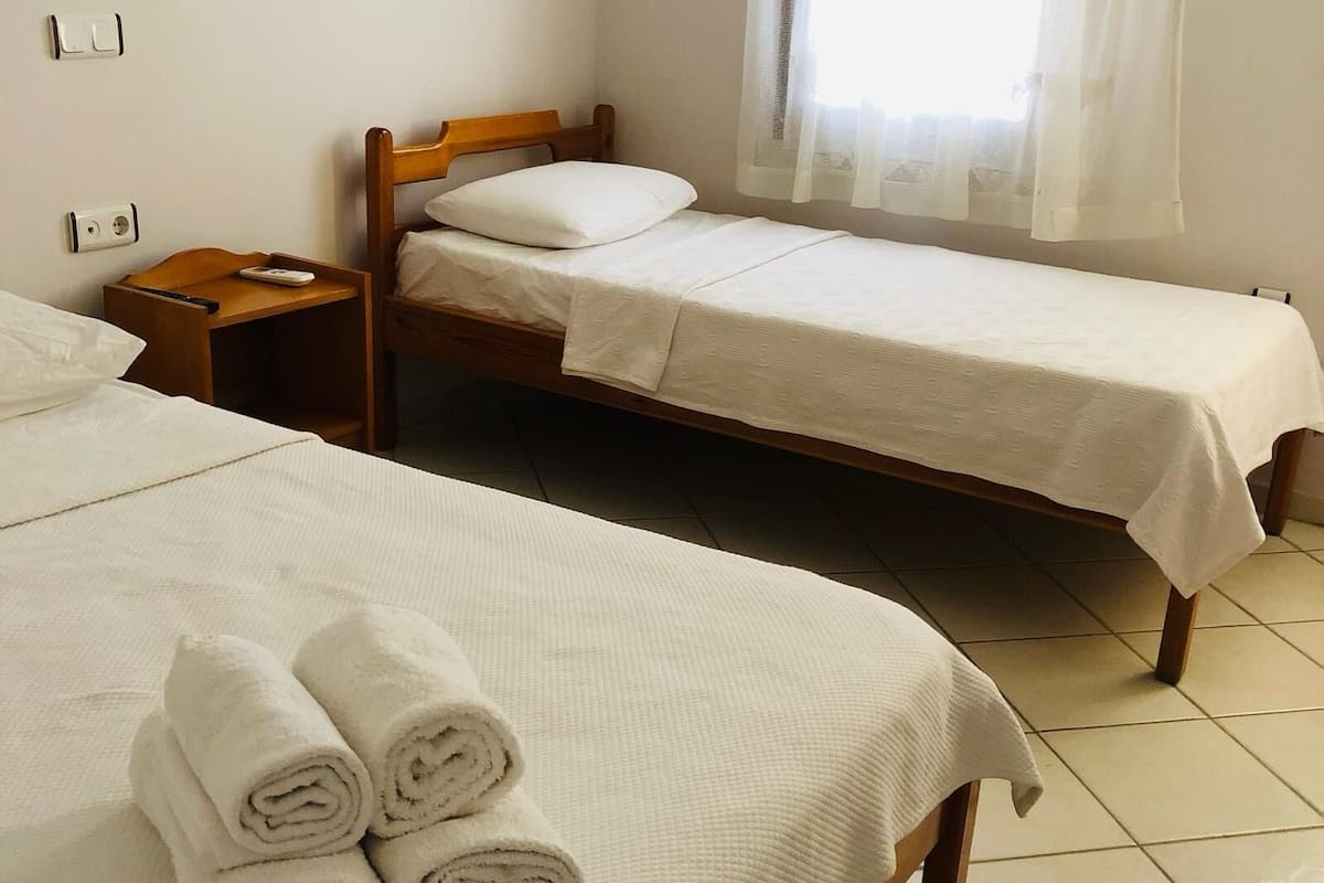 Quarto de hotel com duas camas de solteiro, uma mesinha de cabeceira de madeira e uma janela com cortinas translúcidas.