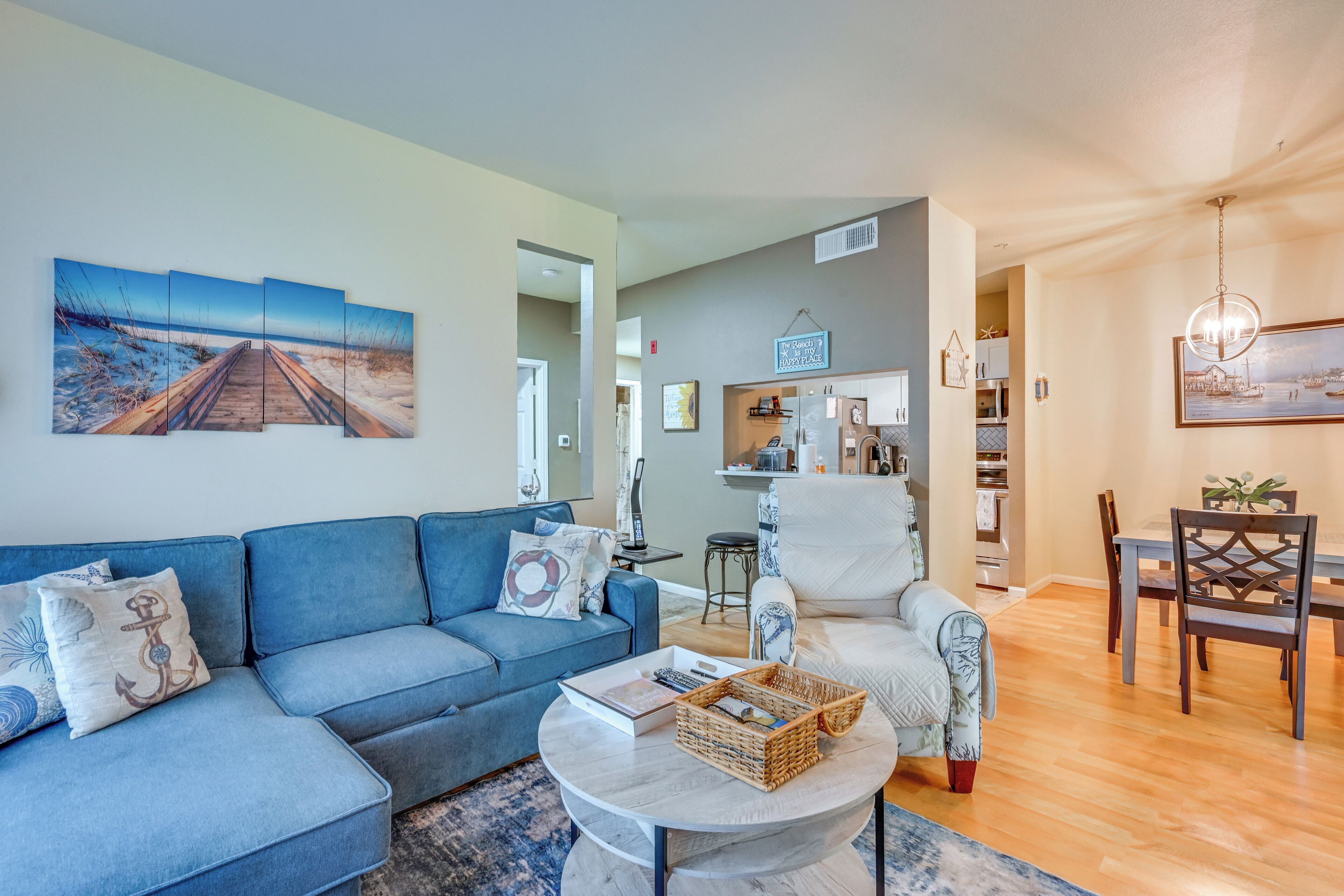 Ein Wohnzimmer mit einem blauen Sofa, einem weißen Sessel, einem Esstisch mit Stühlen und einer Küche im Hintergrund.