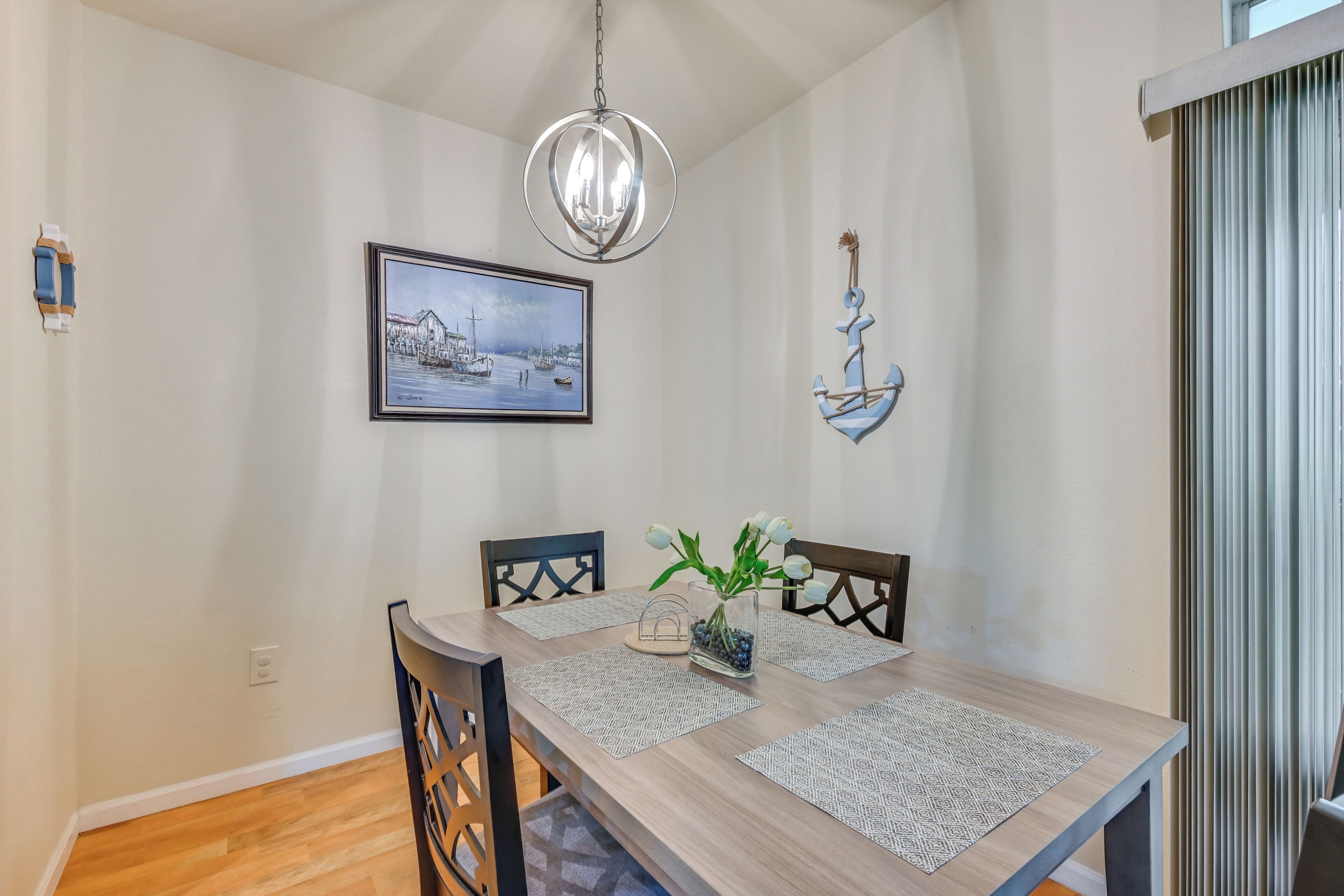 A dining area with a wooden table, four chairs, a chandelier, a framed picture, and a decorative anchor.