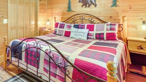 Un dormitorio de cabaña de madera con estructura de cama de metal, colcha acolchada, almohada con la inscripción 'bienvenido a casa', mesita de noche de madera y lámpara de pared.