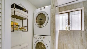 Een kleine wasruimte met twee gestapelde wasmachines, een plank met opgevouwen handdoeken en een raam met decoratief roosterwerk.