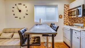 Una cocina compacta con una mesa de comedor, sillas, un reloj, un ventanal con persianas, un microondas y una estufa.