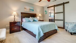 A bedroom with a bed, two bedside tables with lamps, a ceiling fan, a sliding barn door, and a view of the outside.