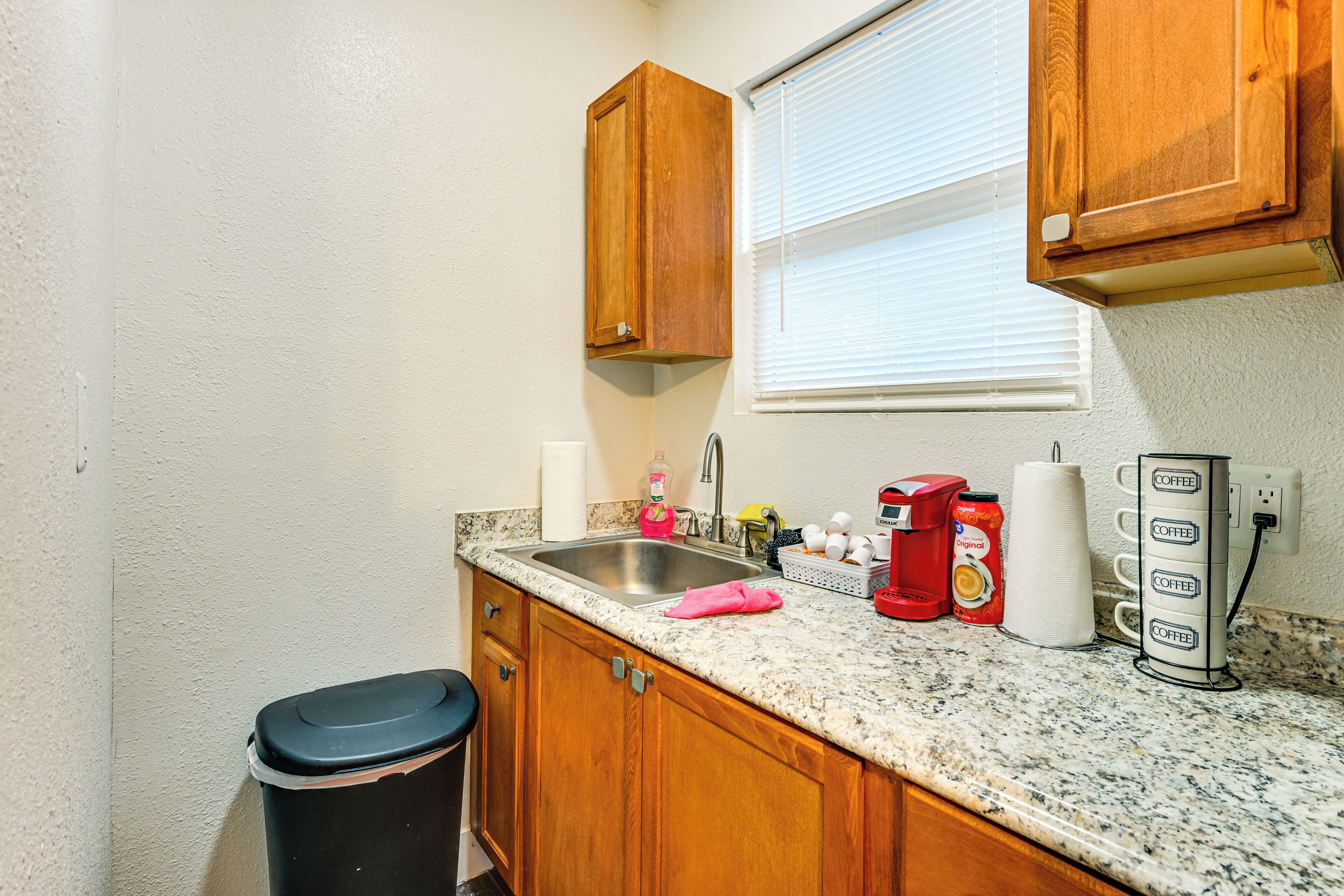 A kitchen with wooden cabinets, a granite countertop, a stainless steel sink, a coffee maker, and a trash can.