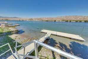 Ein klarer Blick auf einen Fluss mit vertäuten Booten links, einer hölzernen Anlegestelle, die sich ins Wasser erstreckt, und einem Metallgeländer, das zur Anlegestelle führt.