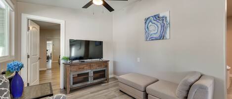 A living room with a ceiling fan, a flat-screen TV on a wooden entertainment unit, a beige sofa, and a patterned armchair.