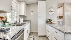 Eine moderne Küche mit weißen Schränken, Granit-Arbeitsplatten und Edelstahlgeräten. An der Wand hängt ein Willkommensschild und auf der Arbeitsfläche steht eine kleine Pflanze.