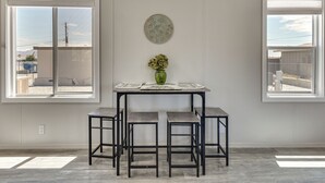 Ein Essbereich mit Tisch und vier Hocker, einer Vase mit Blumen sowie zwei Fenstern mit Blick auf Gebäude.