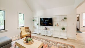 Un salon moderne doté d’un grand téléviseur à écran plat, d’étagères blanches, d’un fauteuil en osier, d’une table basse agrémentée d’objets décoratifs et d’un tapis à motifs.