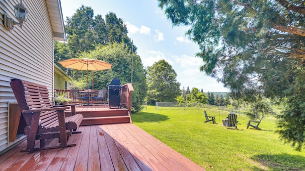Una terraza de madera con mobiliario exterior, una sombrilla y vistas a un césped con árboles.