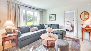 Una sala moderna con un sofá seccional gris, una mesa de centro redonda, un tapete estampado y vistas a un jardín a través de amplios ventanales.