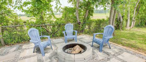 A fire pit surrounded by three blue Adirondack chairs, set on a stone patio with a view of a grassy field and trees.