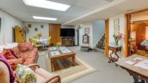 Amplia sala de estar con sofá, sillones, una mesa de centro y televisor. Se observa un mueble de madera y una pequeña mesa con un jarrón de flores.