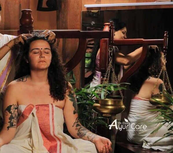 Una persona recibe un tratamiento tradicional, con una olla y una cadena colgante visibles, lo que sugiere un entorno de spa o bienestar.