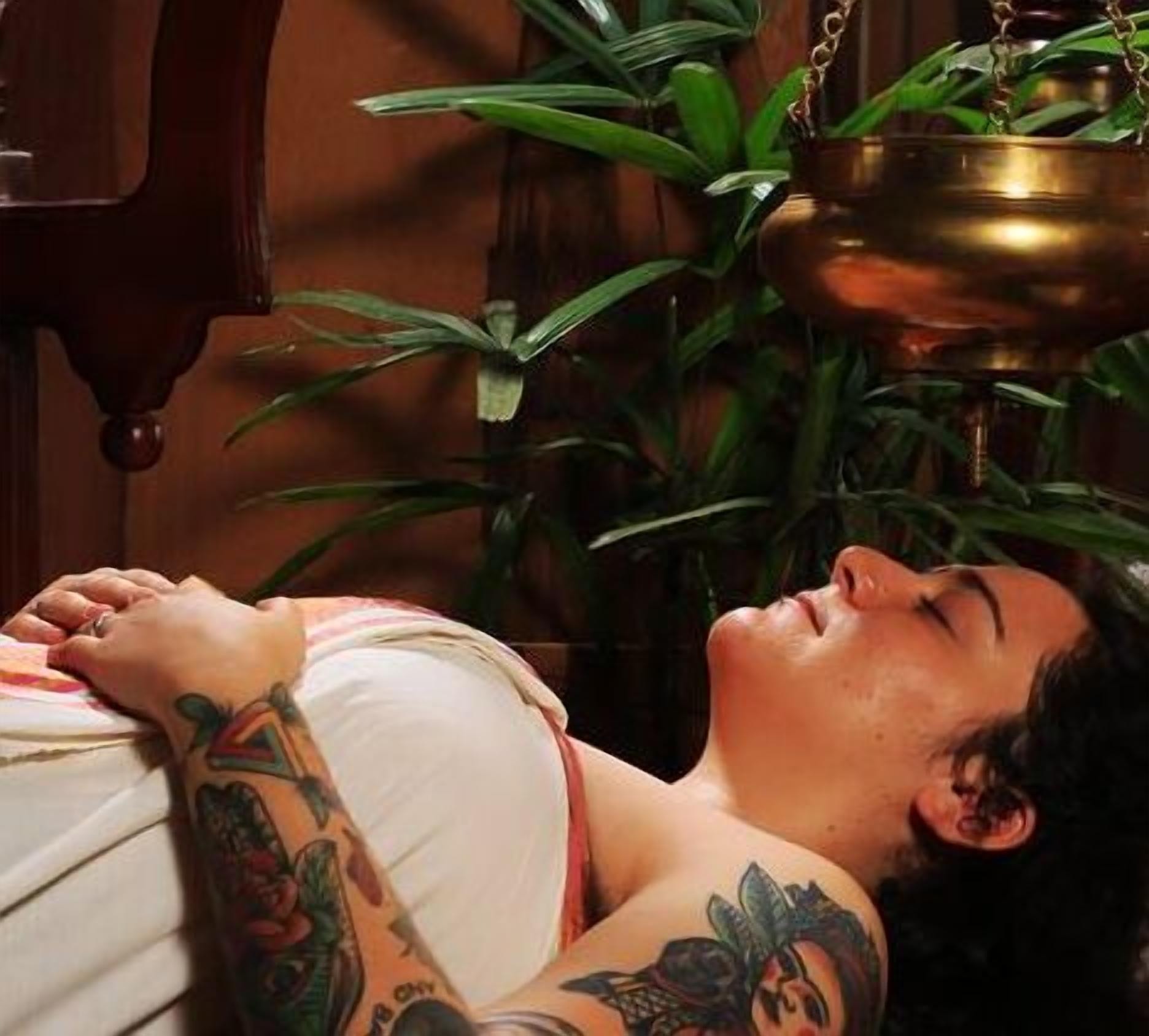 A person lying down with tattoos on their arms, a brass lamp, and green plants in the background.