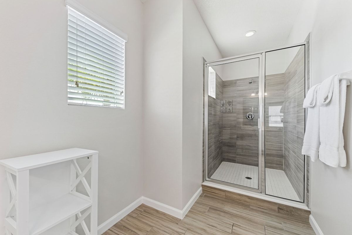 Un baño moderno con ducha acristalada, tocador blanco y ventana con persianas.