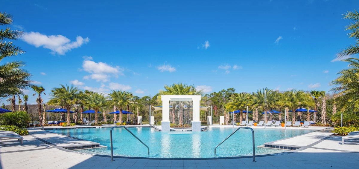 Una piscina con un elemento central de agua, rodeada de palmeras y sombrillas azules.