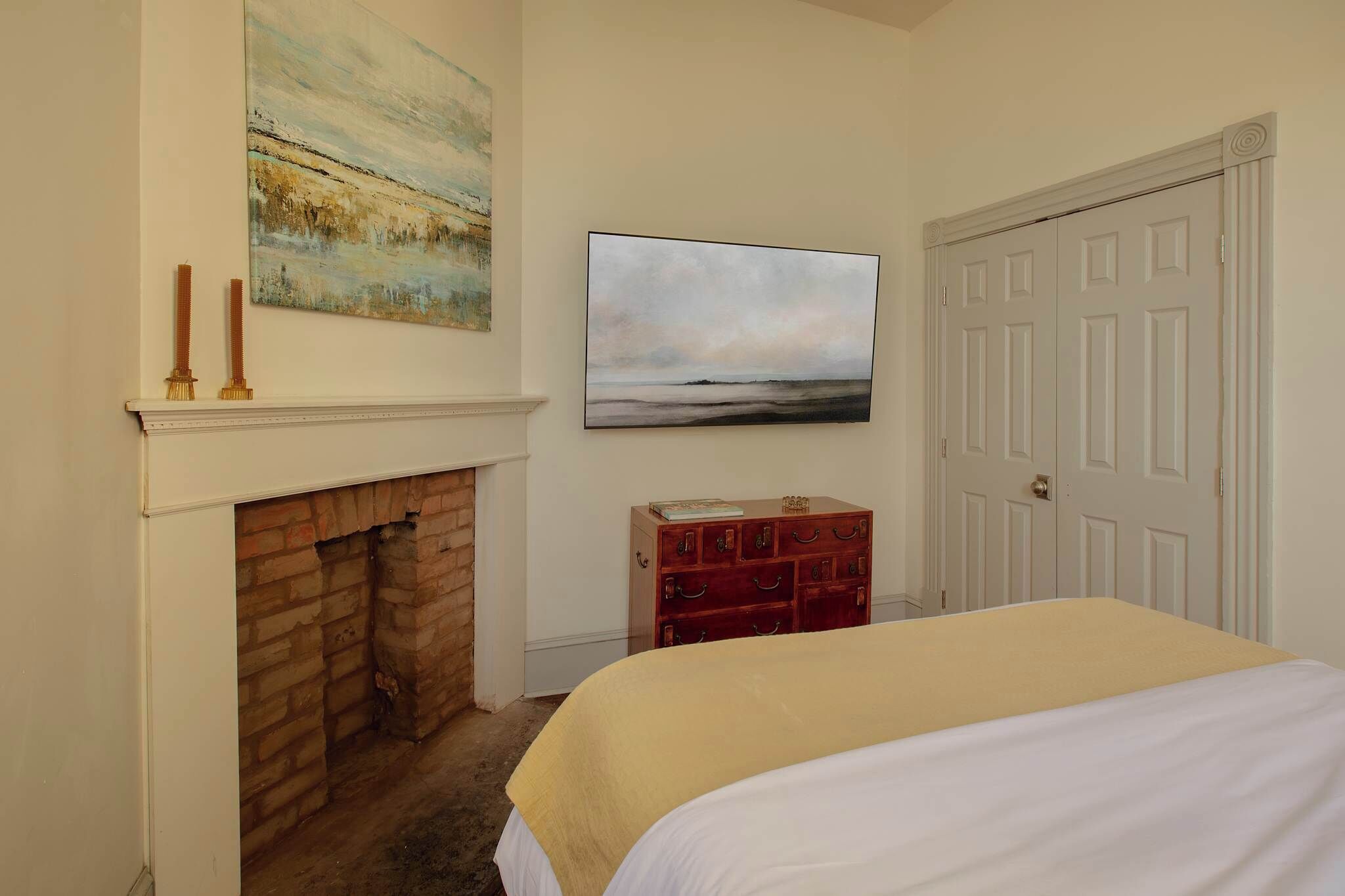 A bedroom with a fireplace, a bed with a yellow bedspread, a red dresser, and two paintings on the wall.