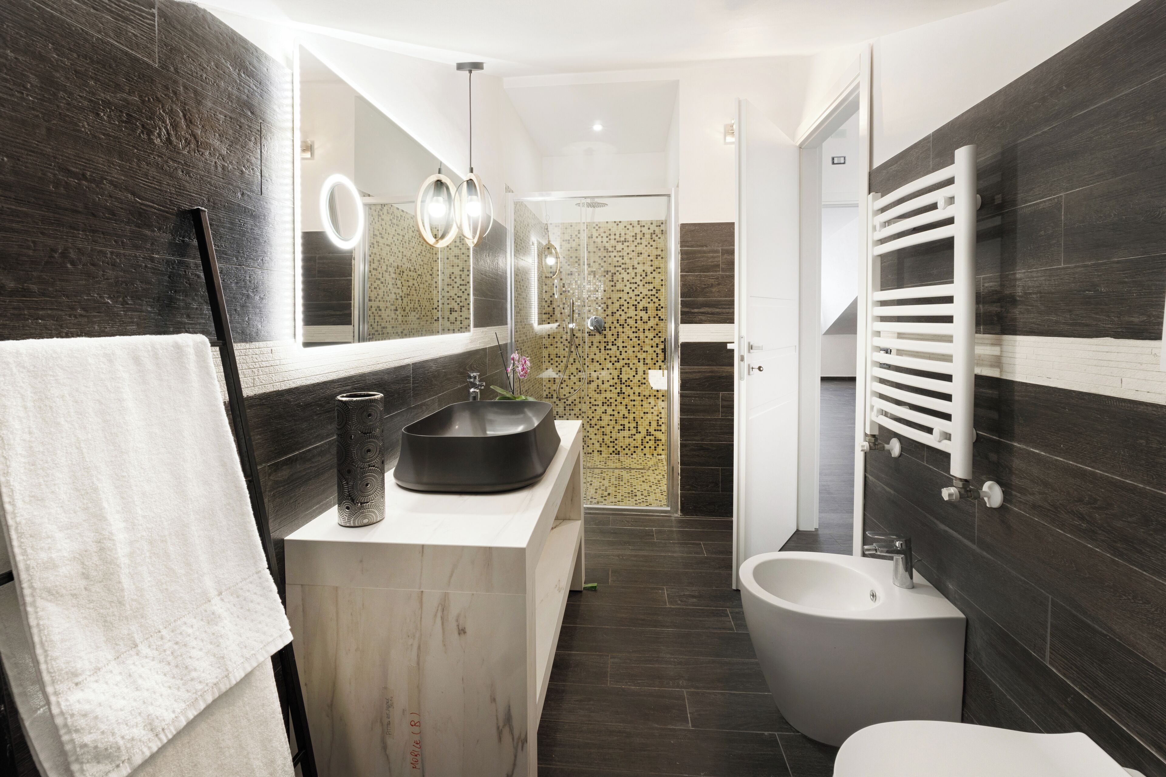 A modern bathroom with a large mirror, a black sink, a bidet, and dark tiled walls.