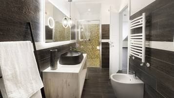 Un baño moderno con un espejo grande, un lavamanos negro, un bidé y paredes de azulejos oscuros.