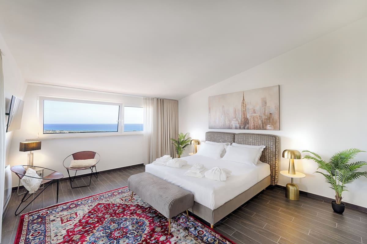 Camera d'albergo con un letto grande, vista sul mare, un dipinto di una città, una scrivania con una lampada e una pianta in vaso.