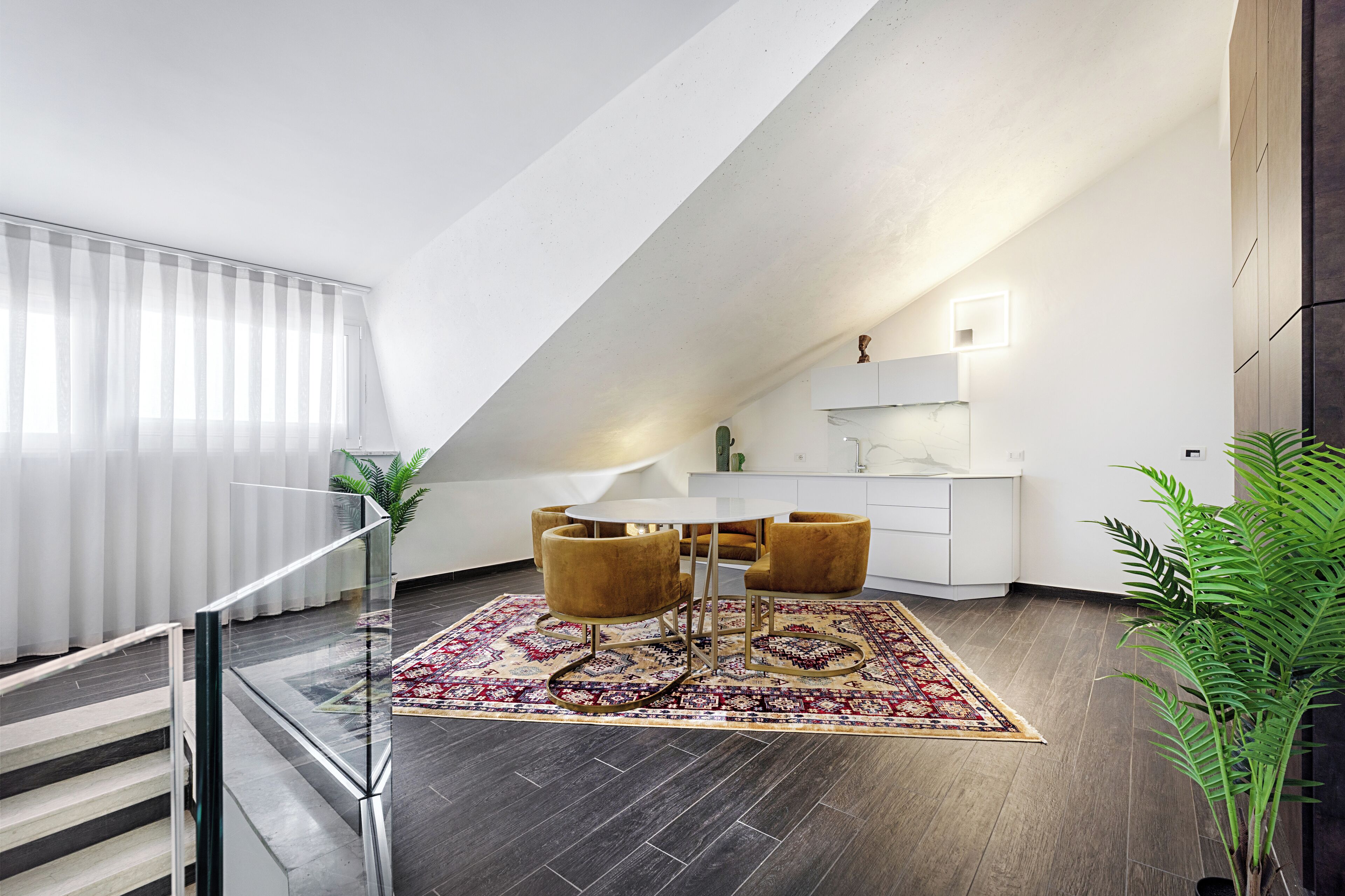 A modern dining area with a round table and chairs, a patterned rug, and a staircase leading to a balcony.
