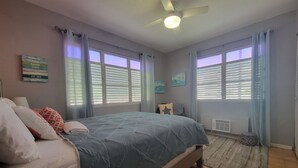 Un dormitorio con cama, ventilador de techo y dos ventanas con cortinas.