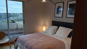 A bedroom with a large bed, a bedside table with a lamp, a chair, and a view through a sliding glass door.