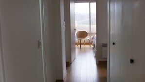 A hallway with a wooden floor leading to a room with a large window, a chair, and a hanging light fixture.