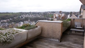 A rooftop terrace with a view of a town, a seating area with a table and chairs, and a planter with flowers.