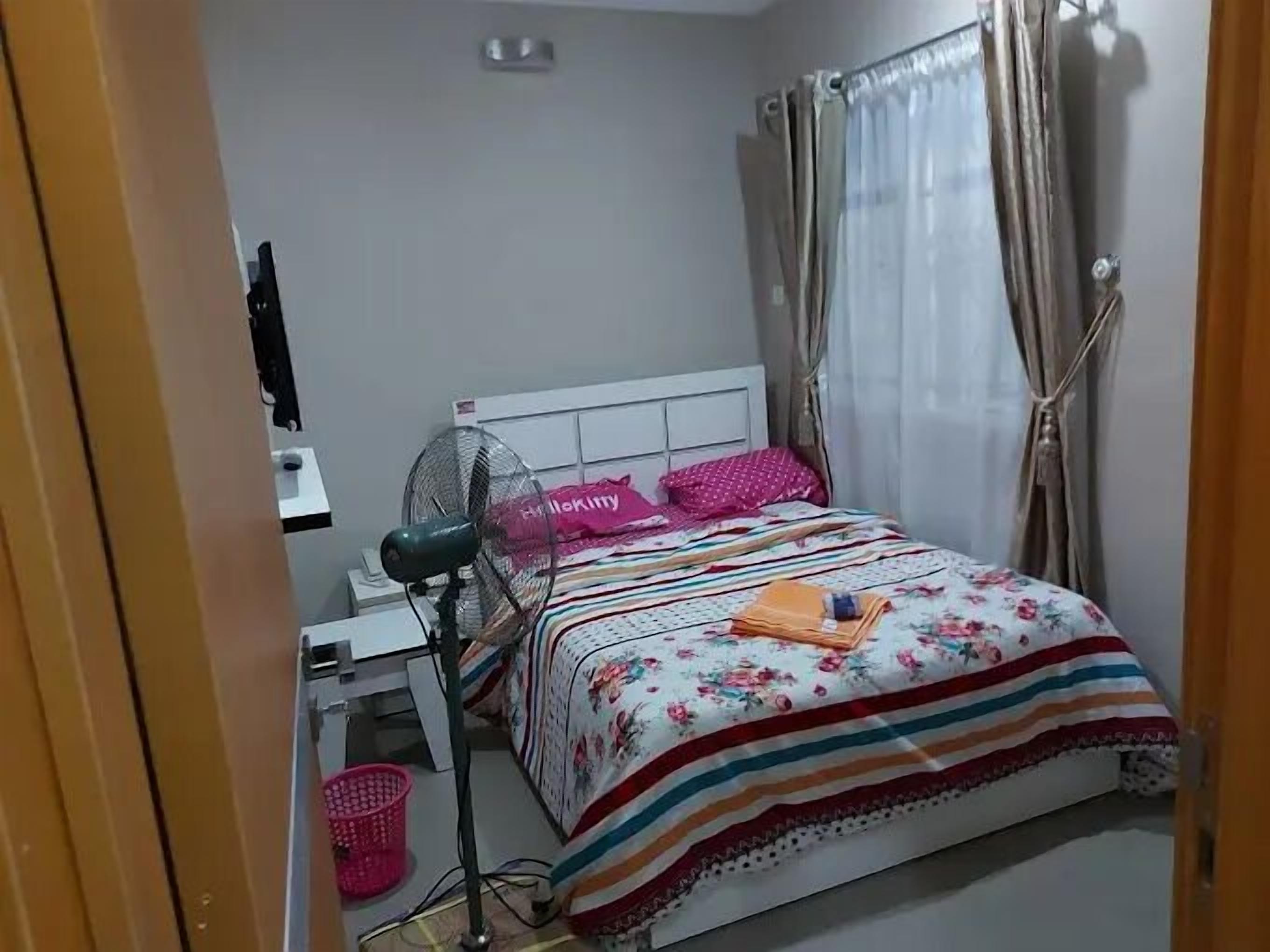 Ein Schlafzimmer mit einem Bett, einem Schreibtisch, einem Stuhl, einem Ventilator und einem Fenster mit Vorhängen.