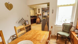 Un coin repas avec une table en bois, des chaises et une assiette décorative. Il y a une fenêtre donnant sur la rue, un réfrigérateur orné d'aimants et une chaise avec un coussin.