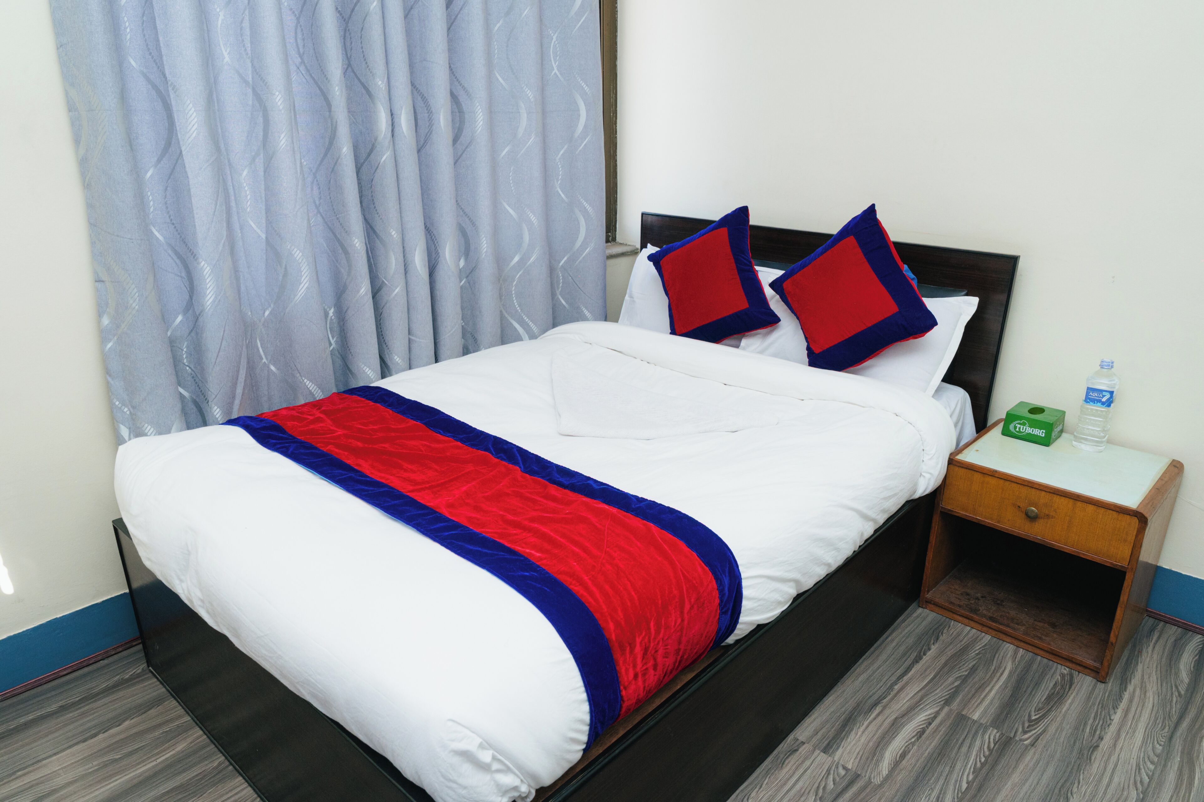 A neatly made bed with a white, red, and blue bedspread, a wooden nightstand with a water bottle, and a curtain on the window.